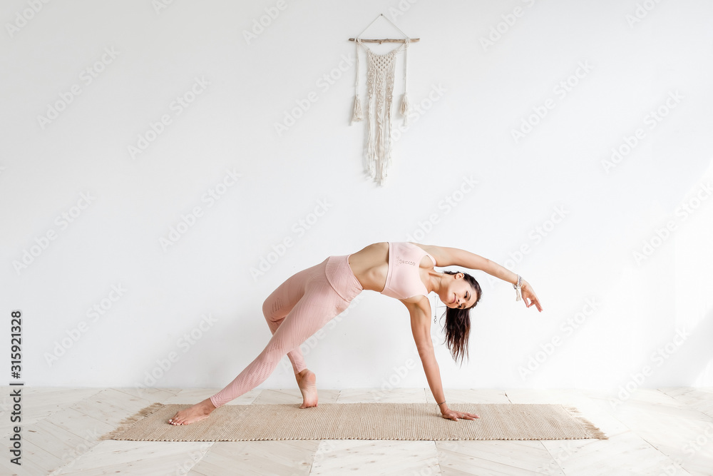 Young sporty attractive woman practicing yoga on wooden floor doing Wild Thing, Flip-the-Dog exercise, Camatkarasana pose. Wearing sportswear pink pants and top, working out, indoor, white yoga studio