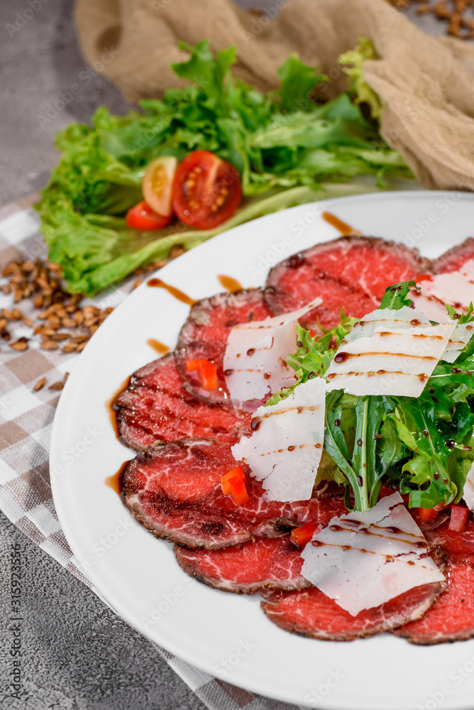 Fresh salad with beef carpaccio, arugula, parmesan, tomatoes and sauce on white plate on light concrete background in beautiful composition with fresh vegetables, napkin and light bread. Close up