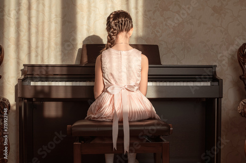 An pretty girl in a pink dress with  a bow is practicing on the piano at home. Back view.