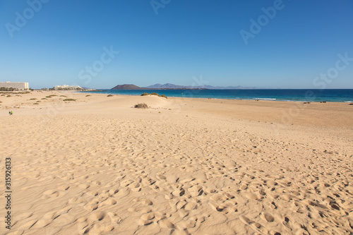 Fototapeta Naklejka Na Ścianę i Meble -  View on sand dune beach from Vuerteventura with Lanzarote and Lobos canary island in the background - golden  yellow sand dunes light blue sky summer season holiday travel vacation seascape mountains