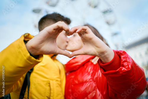 young romantic couple on date, embracing and kissing through a heart gesture made with their fingers, spending time together in the city. Lovers wearning in bright yellow and red down jackets. ferris photo