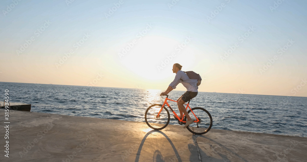 Fototapeta premium Young handsome male in casual wear ride on the colorful bicycle on the morning beach against beautiful sunset and the sea