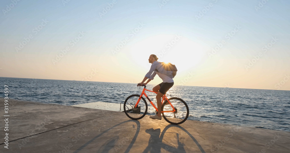Fototapeta premium Young handsome male in casual wear ride on the colorful bicycle on the morning beach against beautiful sunset and the sea