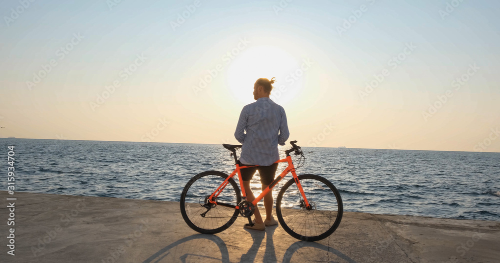 Fototapeta premium Young handsome male in casual wear ride on the colorful bicycle on the morning beach against beautiful sunset and the sea