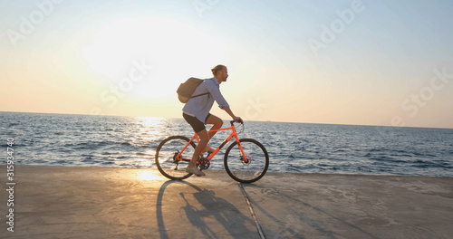 Young handsome male in casual wear ride on the colorful bicycle on the morning beach against beautiful sunset and the sea