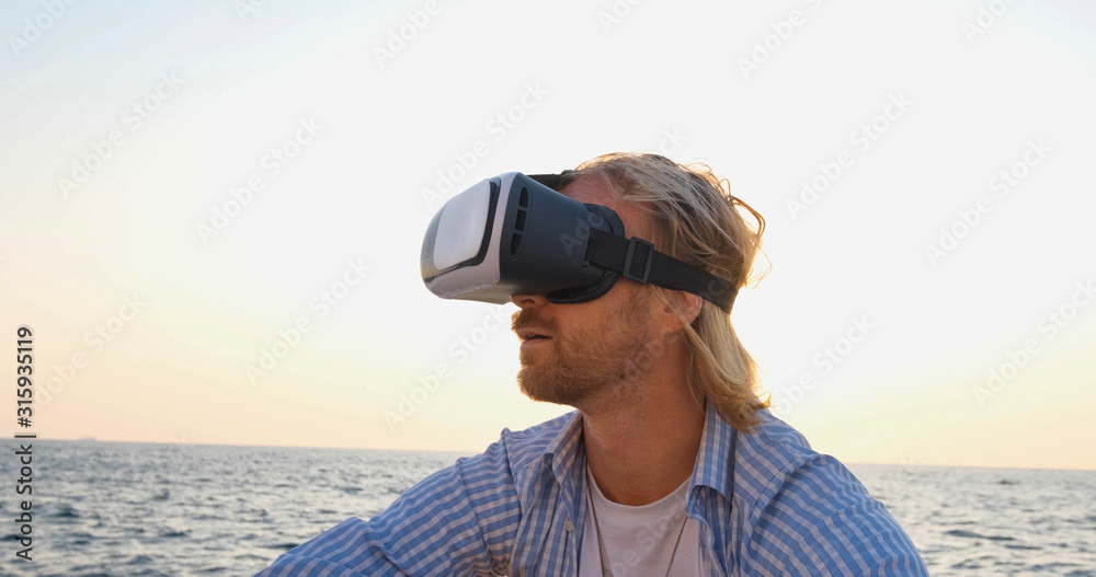 Portrait of young handsome male on the beach wearing virtual reality glasses, beautiful sunrise on the background Portrait of young handsome male on the beach wearing virtual reality glasses, beautiful sunrise on the background