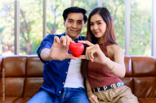 Asian young couple sitting smiling and showing red heart on couch in living room