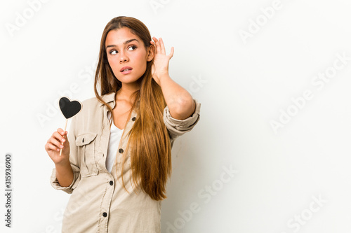 Young caucasian woman holding a heart shape trying to listening a gossip.