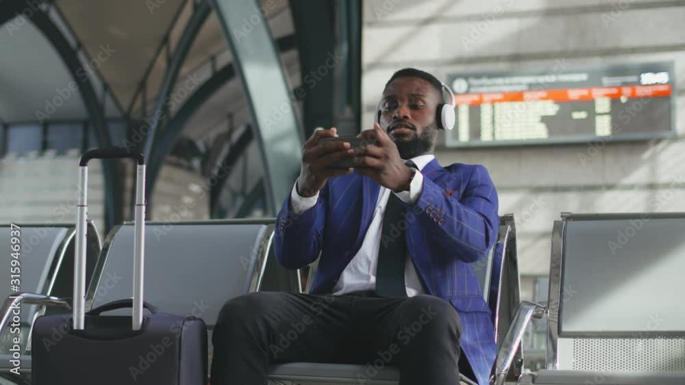 Afro-american man in formal wear holding mobile phone and plays games relaxing while sitting in waiting room of international airport. Businessman waiting for train and using gadgets