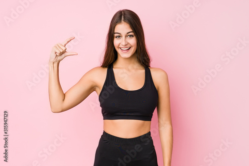 Young caucasian fitness woman doing sport isolated holding something little with forefingers, smiling and confident.