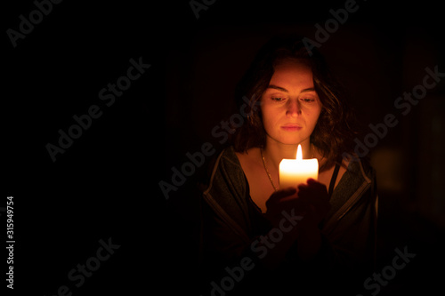 A sad religious orthodox brunette girl with brown eyes looks piercing and sad looking, holding a bright, lighted candle in folded hands. The girl on the right. Horizontal view