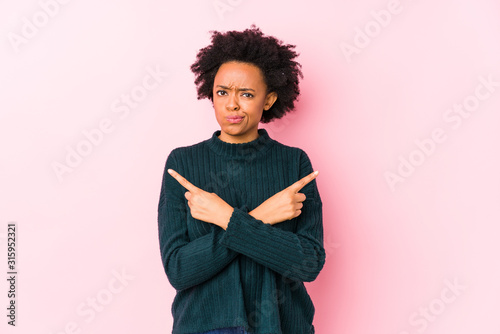Middle aged african american woman against a pink background isolated points sideways, is trying to choose between two options. © Asier