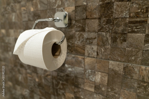 A fresh roll of toilet paper with a folded end hanging on a toilet roll holder against a brown tiled wall