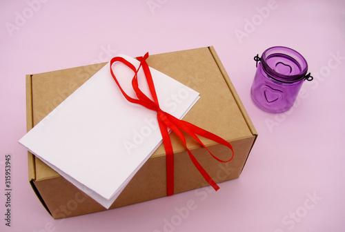 A holiday gift box wrapped with a red bow and a sub-note (empty list) on a pink background . Valentine's day. Mockup.  Purple candlestick with a heart. 