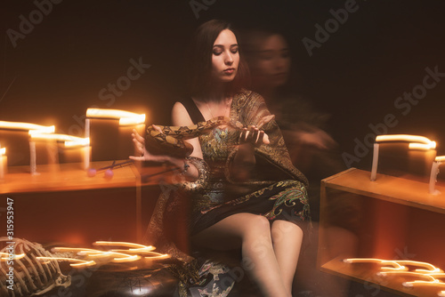 a witch with a snake on her neck and in her hands sits in a haze against a background of candles and a skeleton