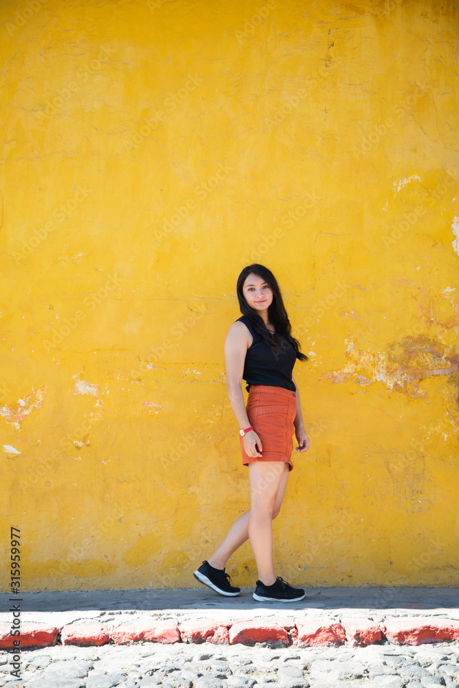 Fototapeta premium Traveling girl in colonial city - Hispanic woman standing in front of yellow old wall - Young woman is posing in the streets of the historical city Guatemala