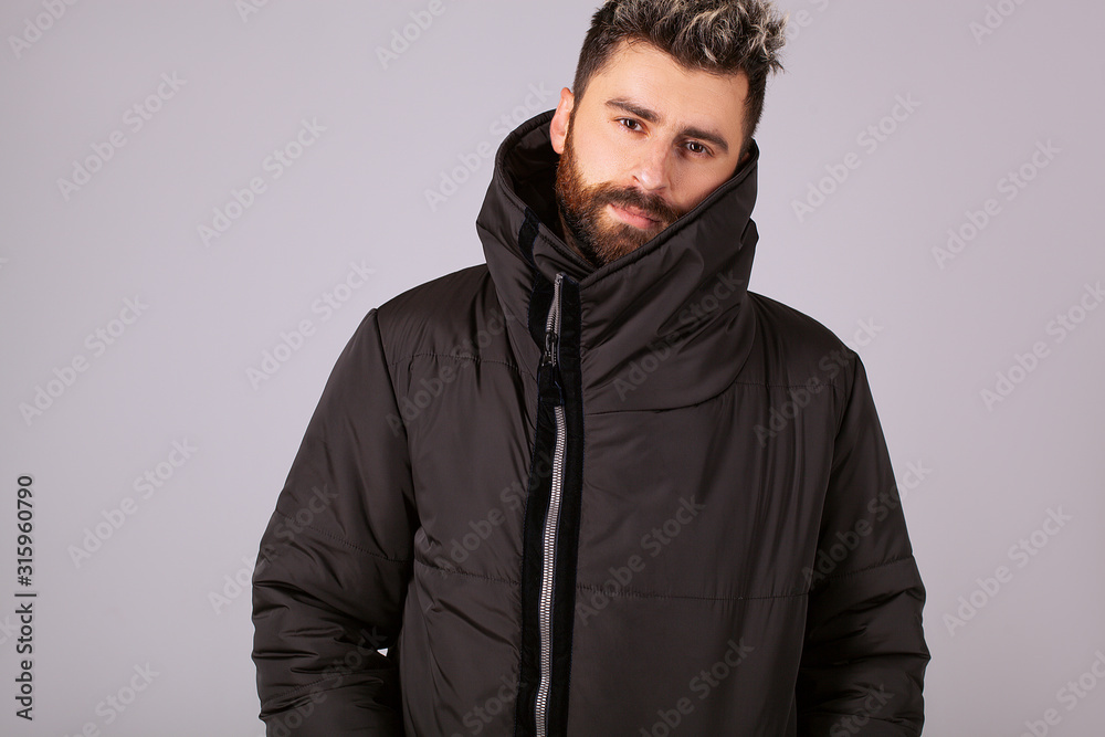 A confident guy in a down jacket is looking at the camera. Warm jacket in black on a gray background. Confident handsome man. Hairstyle with a styling and a stylish beard A confident guy in a down jacket is looking at the camera. Warm jacket in black on a gray background. Confident handsome man. Hairstyle with a styling and a stylish beard