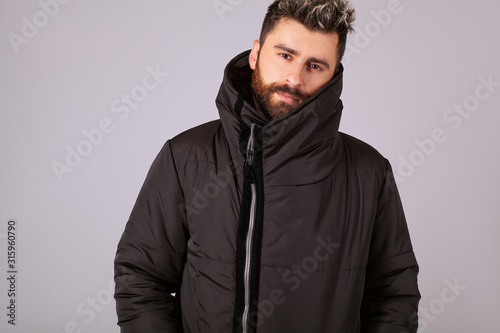 A confident guy in a down jacket is looking at the camera. Warm jacket in black on a gray background. Confident handsome man. Hairstyle with a styling and a stylish beard