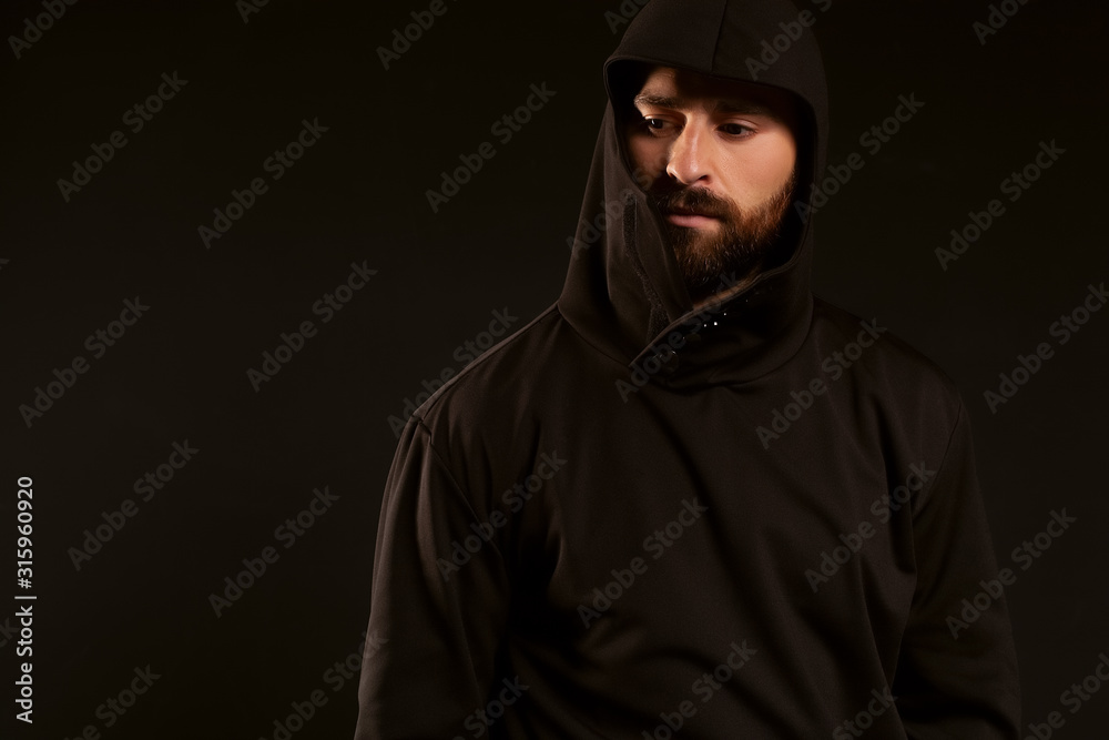Model tests of a man on a black background, wearing a tracksuit. The gaze is lowered to the bottom of the frame. A beautiful beard and mustache on the face, and the head is covered with a hood Model tests of a man on a black background, wearing a tracksuit. The gaze is lowered to the bottom of the frame. A beautiful beard and mustache on the face, and the head is covered with a hood