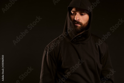 Model tests of a man on a black background, wearing a tracksuit. The gaze is lowered to the bottom of the frame. A beautiful beard and mustache on the face, and the head is covered with a hood