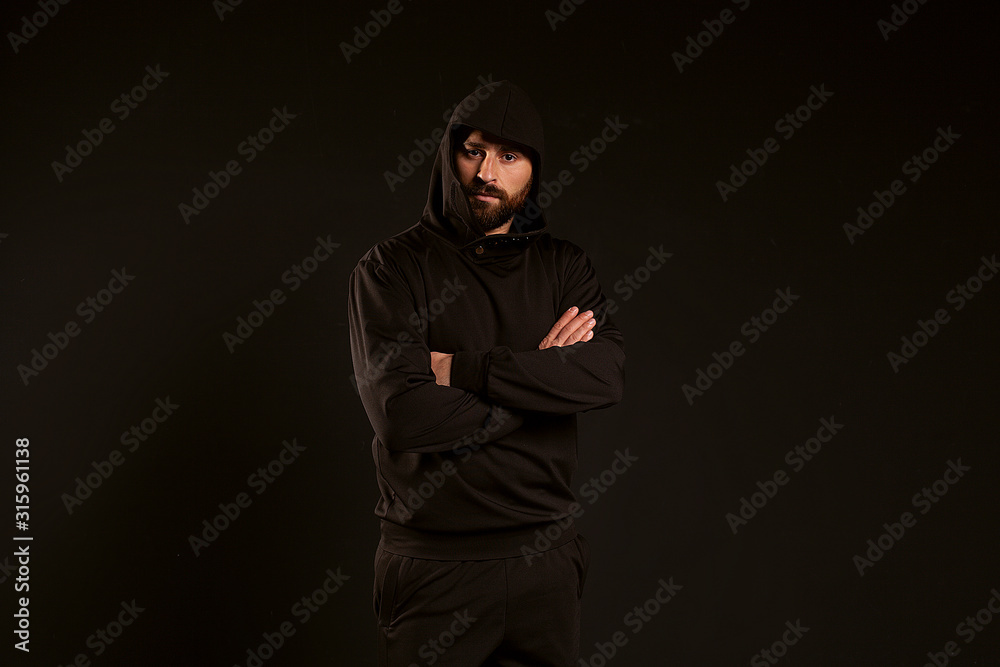 handsome and confident man on a black background. The guy in the black tracksuit folded his arms. Confident look of a sports guy with a beautiful hairstyle and a hood on his head handsome and confident man on a black background. The guy in the black tracksuit folded his arms. Confident look of a sports guy with a beautiful hairstyle and a hood on his head