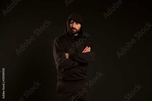 handsome and confident man on a black background. The guy in the black tracksuit folded his arms. Confident look of a sports guy with a beautiful hairstyle and a hood on his head