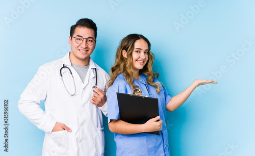 Young doctor couple posing in a blue background isolated holding a copy space on a palm.