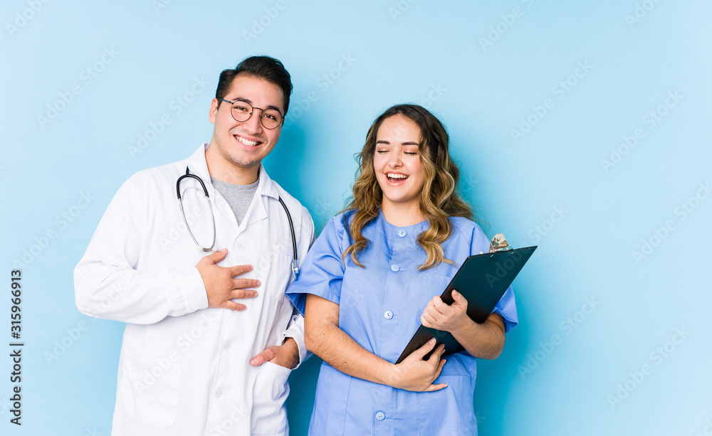 Young doctor couple posing in a blue background isolated laughs out loudly keeping hand on chest.
