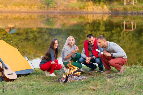 travel, tourism, hike, picnic and people concept - group of happy friends frying sausages on campfire near lake.
