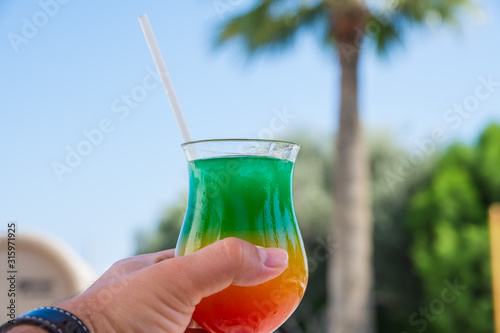 Vacation and rest concept. A man holds in his hand glass with colorful cocktail with drinking straw over the palm trees. Selective focus.