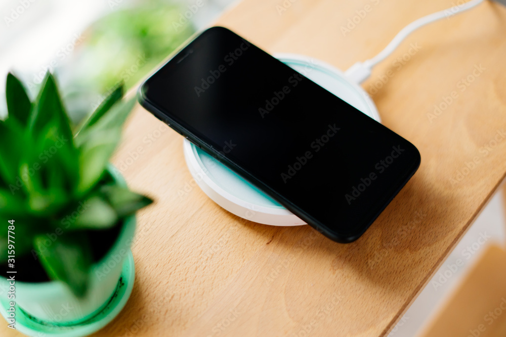 Smartphone in mint silicone case is charged from a wireless charger. The mobile phone is charged on a wooden nightstand or shelf. Smartphone in mint silicone case is charged from a wireless charger. The mobile phone is charged on a wooden nightstand or shelf.
