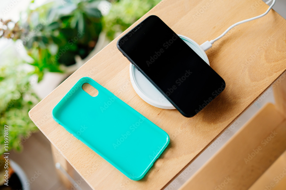 Black smartphone and mint silicone case is charged from a wireless charger. The mobile phone is charged on a wooden nightstand or shelf. Black smartphone and mint silicone case is charged from a wireless charger. The mobile phone is charged on a wooden nightstand or shelf.