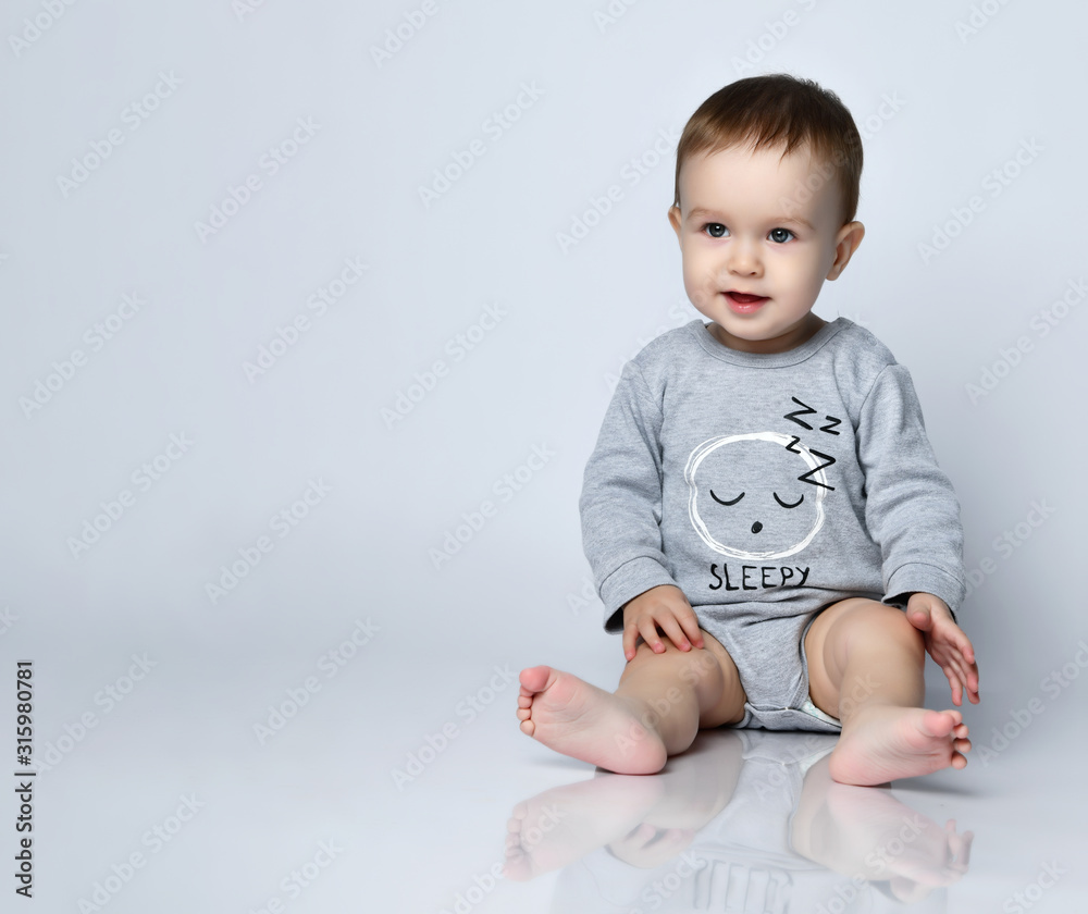 Little baby boy toddler in grey casual jumpsuit and barefoot sitting on floor and smiling over white wall background Little baby boy toddler in grey casual jumpsuit and barefoot sitting on floor and smiling over white wall background