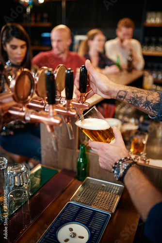 Tattooed caucasian barman pouring beer while standing in pub. Selective focus on hand.