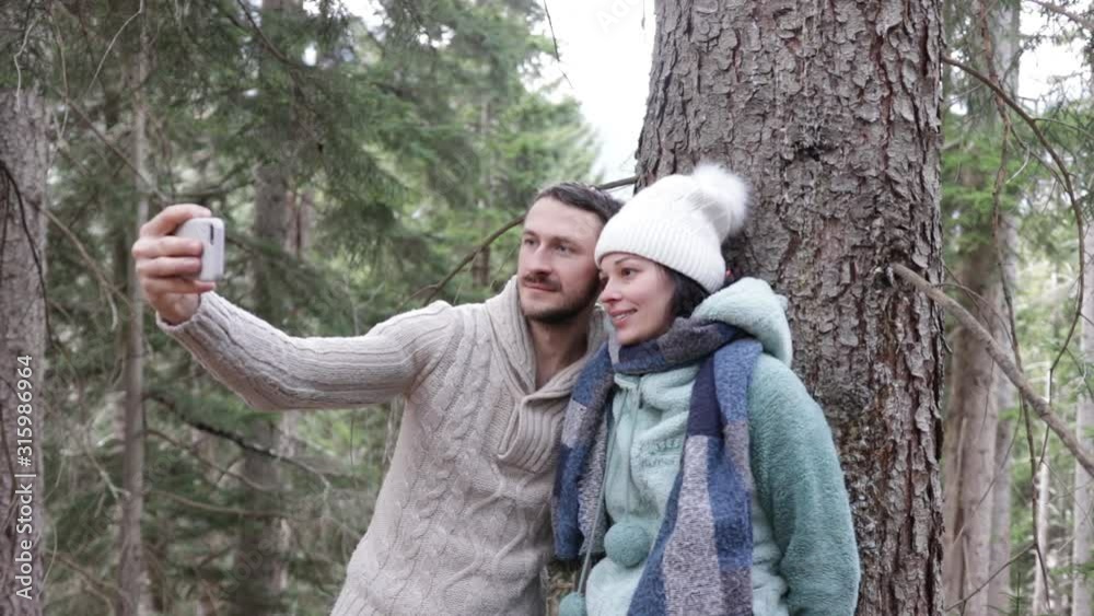 Young married couple in bright clothes adjust the camera and take selfies on the phone or smartphone in the winter forest