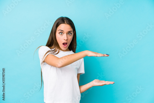 Young caucasian woman  isolated shocked and amazed holding a copy space between hands.
