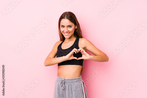 Young caucasian fitness woman posing in a pink background smiling and showing a heart shape with hands.