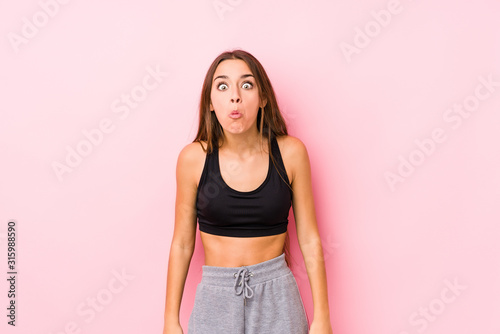 Young caucasian fitness woman posing in a pink background shrugs shoulders and open eyes confused. © Asier