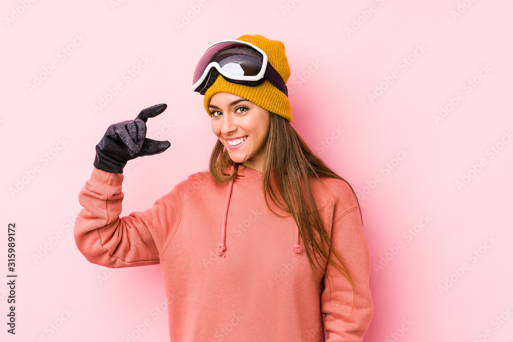 Fototapeta premium Young caucasian woman wearing a ski clothes isolated holding something little with forefingers, smiling and confident.