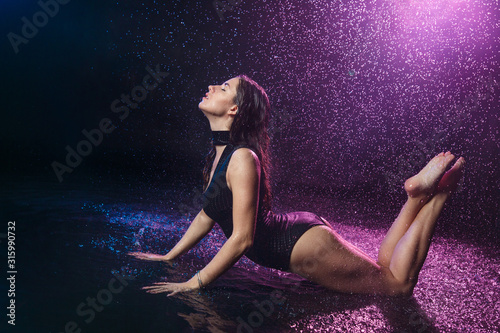 A beautiful sexy girl in a swimsuit lies under the drops of falling water with purple backlight on a black background.