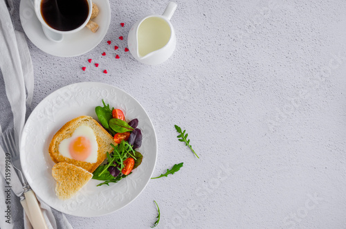 Piece of bread toast cut in shape of heart with egg on a white plate with arugula and cherry tomatoes. Romantic breakfast for Valentine's Day. Top view. Copy space. Horizontal orientation.