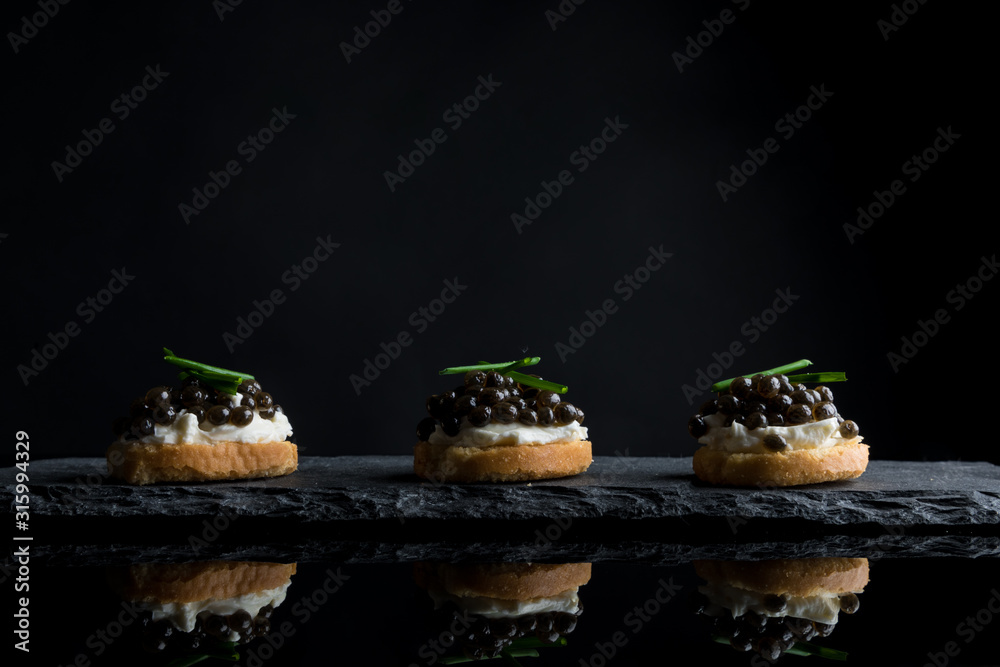 Caviar, three snacks of black caviar on bread and stone plate isolated on black background with reflections, copy space - obrazy, fototapety, plakaty 