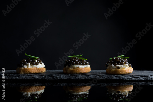 Caviar, three snacks of black caviar on bread and stone plate isolated on black background with reflections, copy space photo