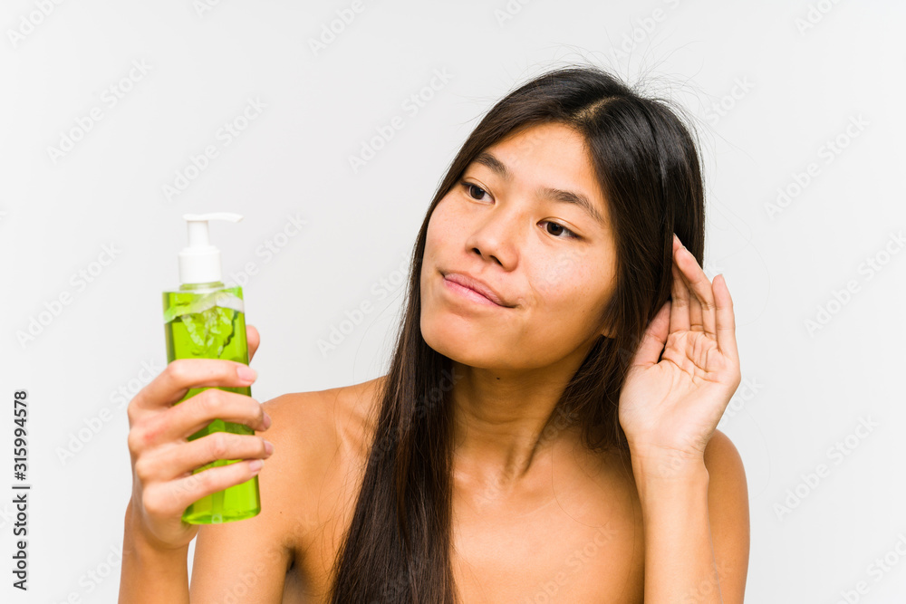 Young chinese woman holding a moisturizer with aloe vera isolated trying to listening a gossip. Young chinese woman holding a moisturizer with aloe vera isolated trying to listening a gossip.