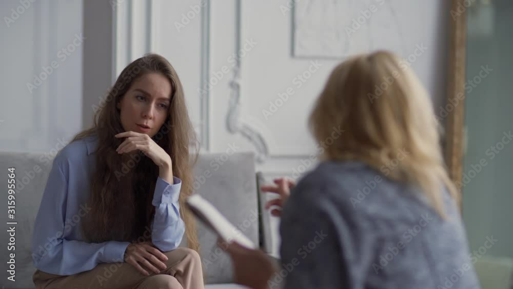 Tracking left shot of worried young woman covering face with hand talking emotionally about problems with therapist holding pen and notepad Tracking left shot of worried young woman covering face with hand talking emotionally about problems with therapist holding pen and notepad