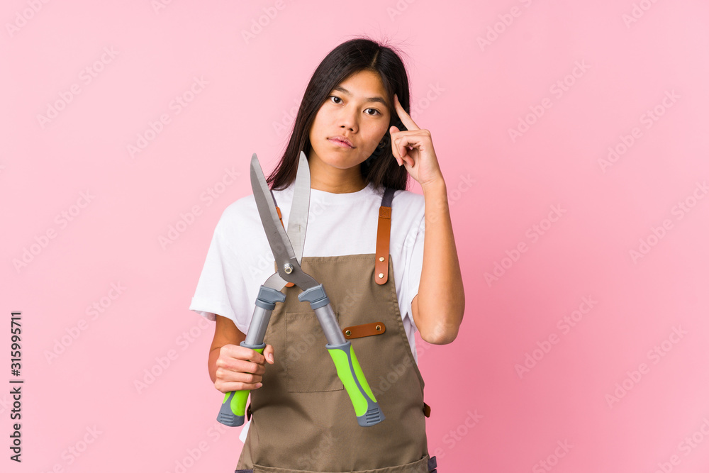 Young chinese gardener woman holding a scissors isolated pointing temple with finger, thinking, focused on a task. Young chinese gardener woman holding a scissors isolated pointing temple with finger, thinking, focused on a task.