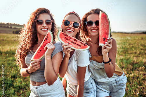 Three young attractive women friends at sunshine are eating watermelon and smiling.