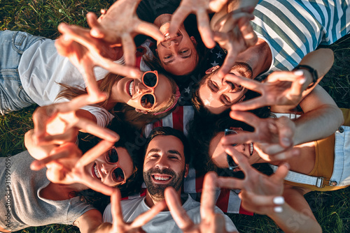 Group of smiling friends lying on grass in circle and showing victory sign outdoors