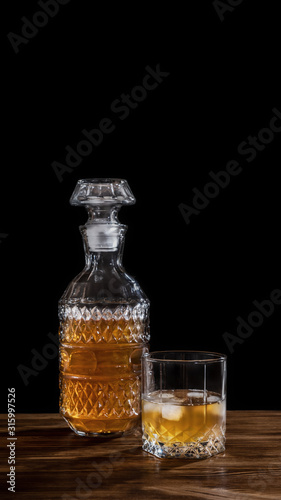 Glass of whiskey with ice and a square decanter isolated on black background on wooden table. Alcohol concept