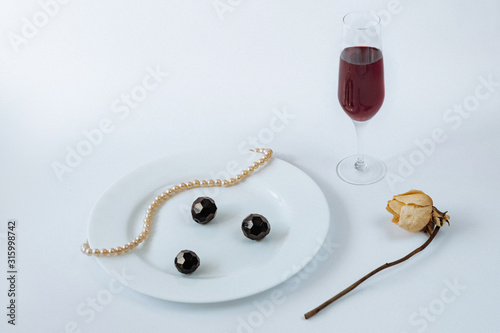 White plate with necklace and black crystals on it, fork, knife, dried rose and glass of champagne on a white background
